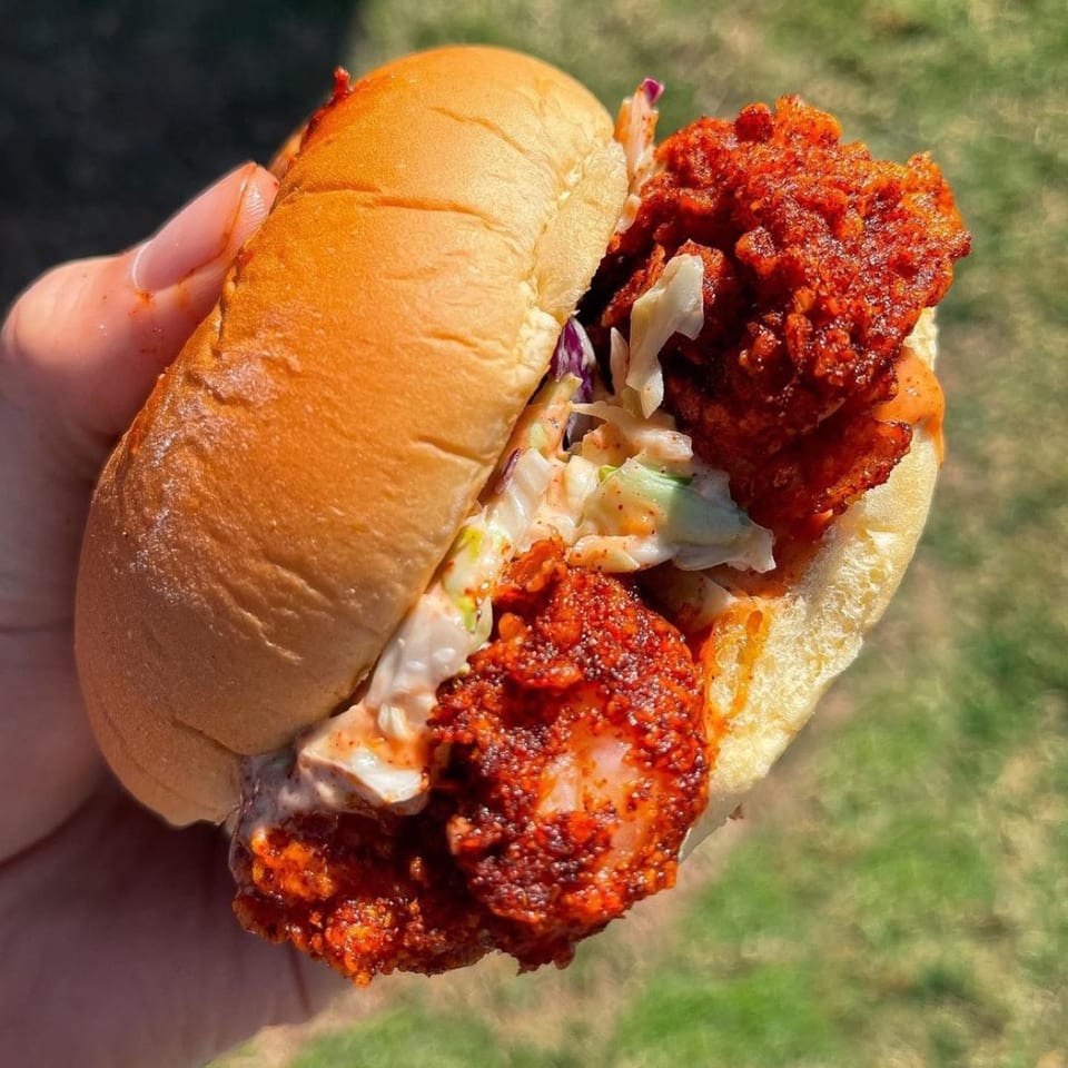 Hand holding a spicy fried chicken sandwich with coleslaw on a brioche bun, outdoors.