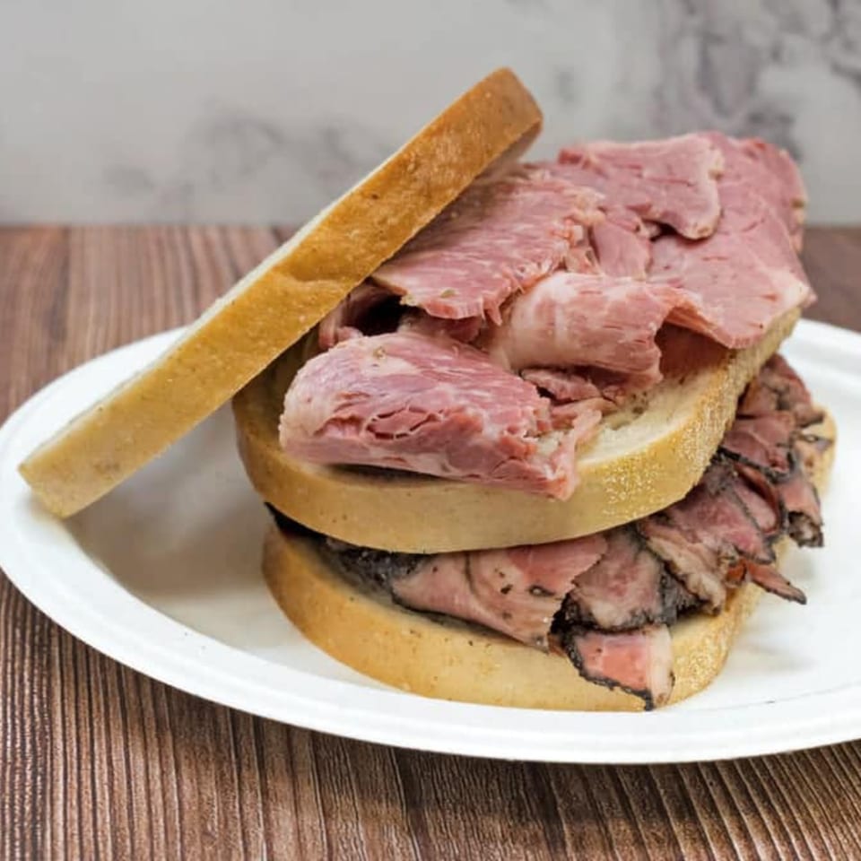 A stacked NY Style Delicatessen sandwich with pastrami and corned beef on white bread.