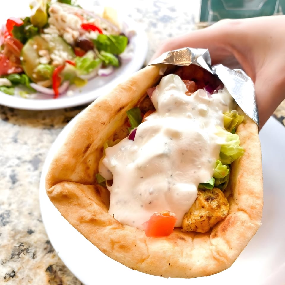 Hand holding a NY Style Delicatessen gyro with white sauce, lettuce, and chicken, with a salad in the background.